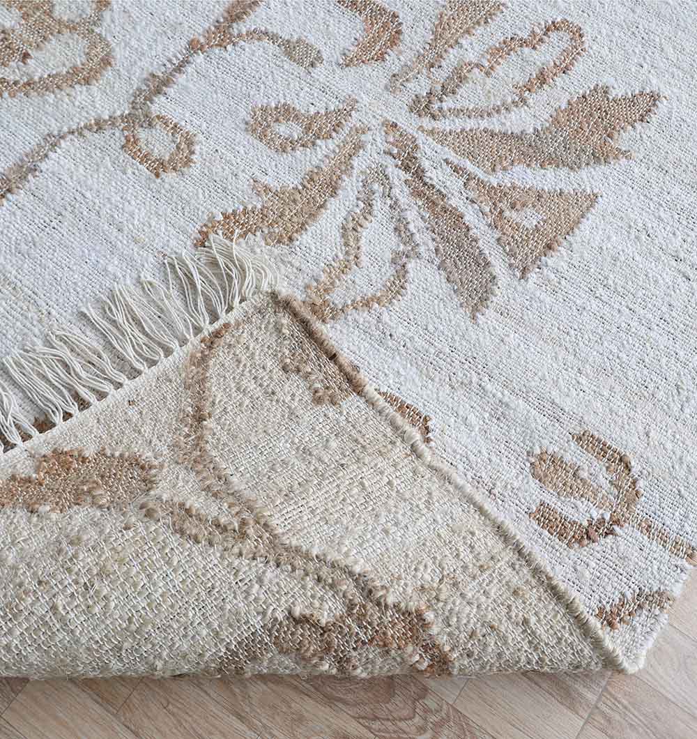 A close-up of the RAMSHA HOME Kilim Rug - Natural Handmade Flatweave Carpet (KLM 022), featuring beige and taupe floral patterns with fringe, partially folded to show the underside, placed on a light wood floor.