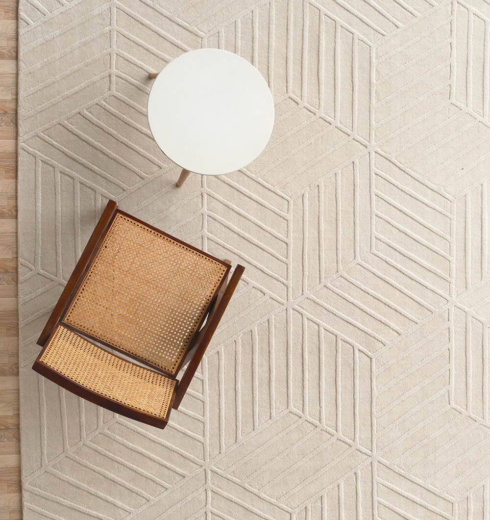 A top-down view of a wooden chair with a woven cane seat beside a small round side table, all placed on the RAMSHA HOME Handmade Off White Decor Rug - LKH 010 (300x400cm) featuring beige geometric patterns.