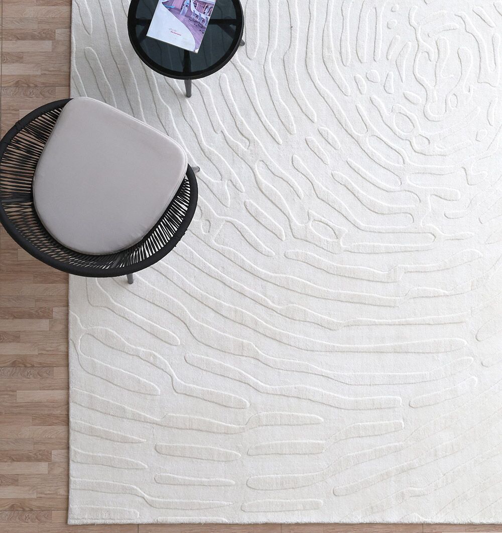 A modern, minimalist room with the RAMSHA HOME Natural Color Home Decor Area Rug - LKH 011, abstract raised patterns, a round black chair with light cushion, and a small black side table with magazines on a light wood floor.