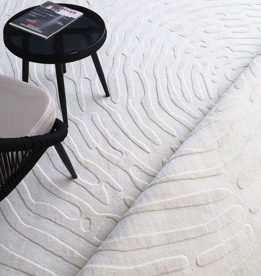 The RAMSHA HOME Natural Color Home Decor Area Rug - LKH 011, featuring wavy, raised patterns, is partially rolled. Beside it are a black round side table with a magazine and a black chair with a light cushion.