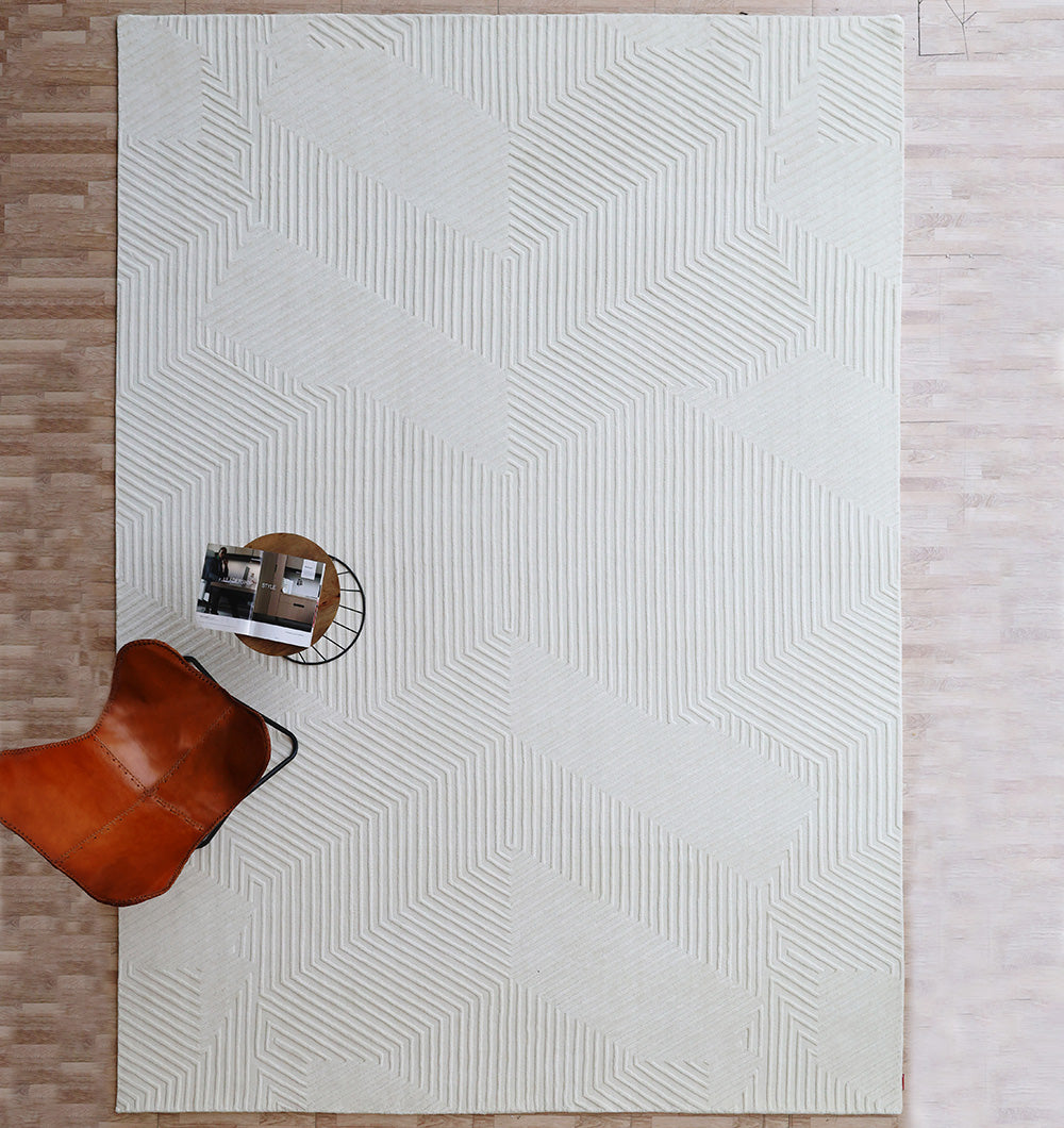 The RAMSHA HOME Handmade Decor Area Rug - LKH 012 in cream covers a light wood floor. In the lower left, a brown leather chair and a small round side table with books rest on the rug, as seen from above.