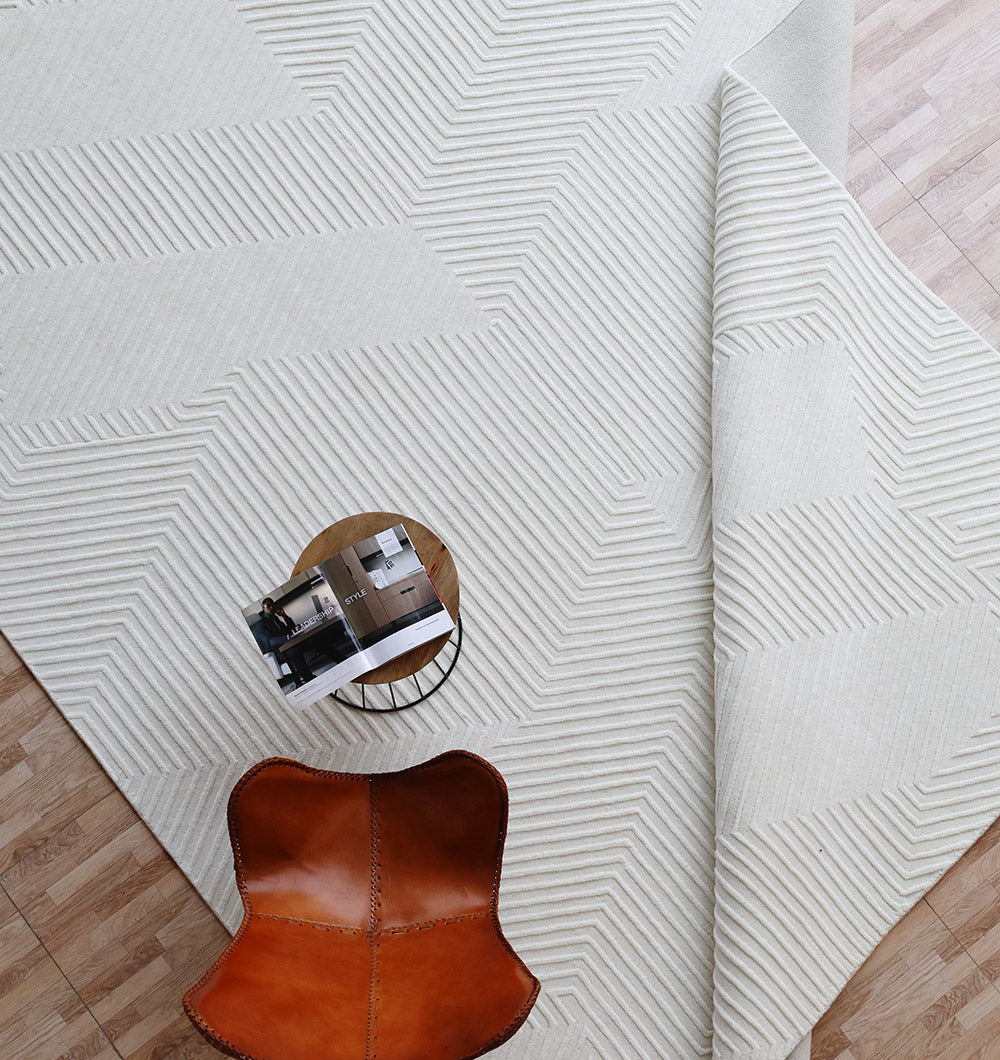 The RAMSHA HOME Handmade Decor Area Rug - LKH 012, featuring a textured ivory finish and geometric patterns, is partially folded on a light wood floor next to a round wooden side table with a magazine and a brown leather chair.