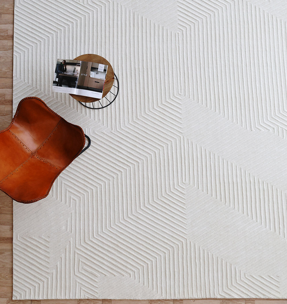 A tan leather chair, a small round table with magazines, and the RAMSHA HOME Handmade Decor Area Rug - LKH 012 in ivory with a textured geometric pattern are seen on a light wood floor.