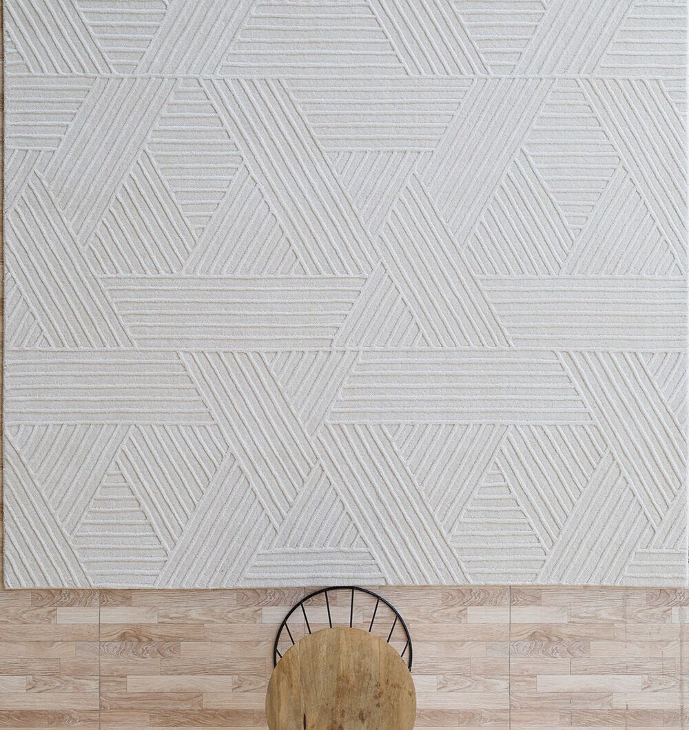 A top-down view of the RAMSHA HOME Off White Color Handmade Area Rug - LKH-013 (200x300 cm) on a wooden floor, with part of a round wooden table and a black metal chair visible at the bottom edge.