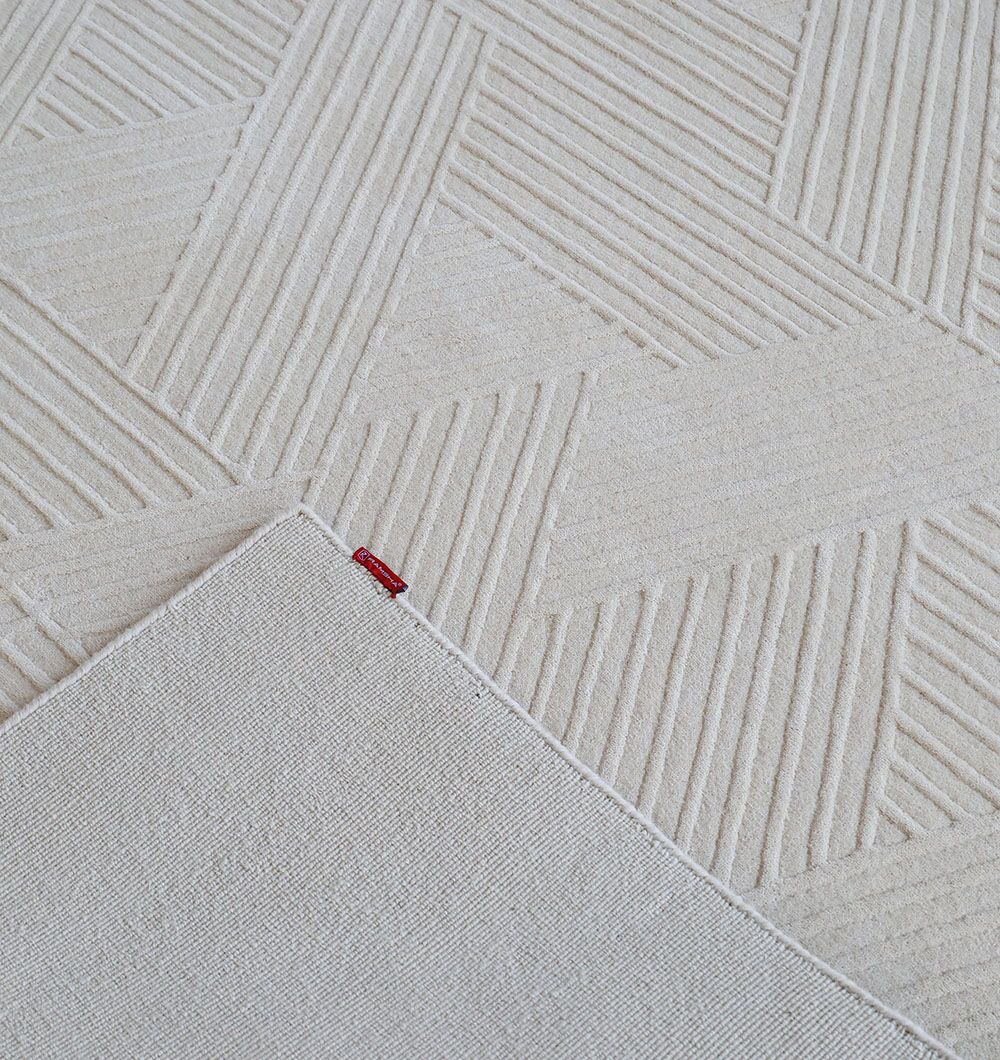A close-up of the Off White Color Handmade Area Rug - LKH-013 (200x300 cm) by RAMSHA HOME shows its textured geometric pattern. One corner is folded, exposing the plain underside and a small red fabric tag on this loom-knotted rug.