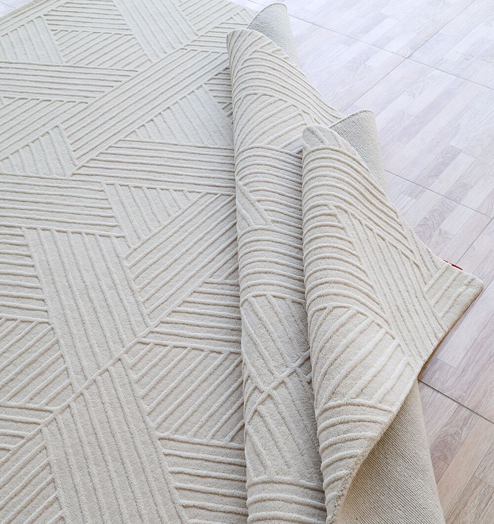 A close-up of the Off White Color Handmade Area Rug - LKH-013 (200x300 cm) by RAMSHA HOME, featuring a loom-knotted texture and subtle beige pattern, partially folded on a light wood floor.
