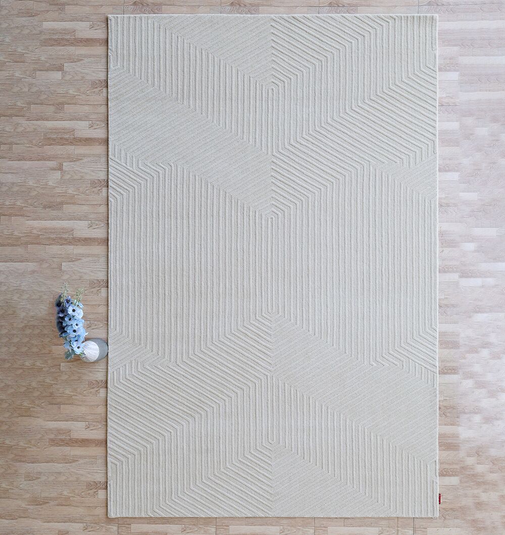The RAMSHA HOME Handmade Area Rug (LKH-014, 200x300 cm) in beige with a geometric pattern lies on a light wooden floor, accented by a small glass vase of purple flowers and a round white dish in one corner.