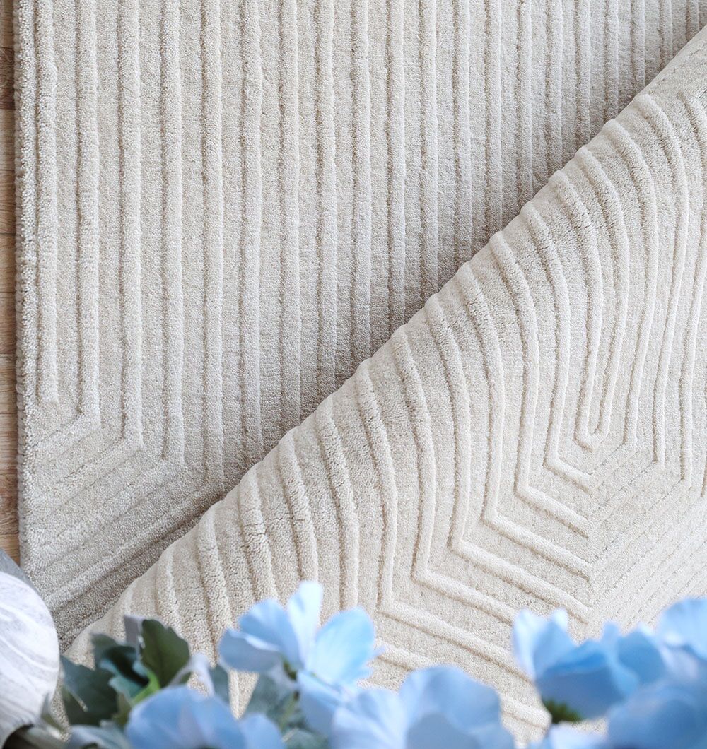 A close-up of the RAMSHA HOME Home Decor Handmade Area Rug - LKH-014 (200x300 cm), featuring textured beige geometric patterns and raised stripes. The folded corner reveals the soft rug, with blue artificial flowers in the foreground.