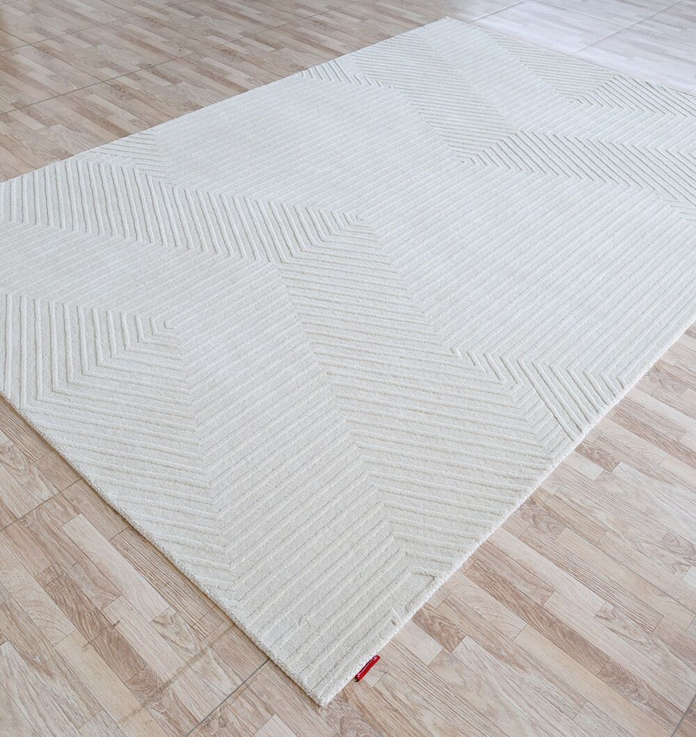The RAMSHA HOME Handmade Area Rug - LKH-014 (200x300 cm) in light beige features a subtle geometric chevron pattern and textured, raised design, accented by a small red tag in one corner on a light wood floor.