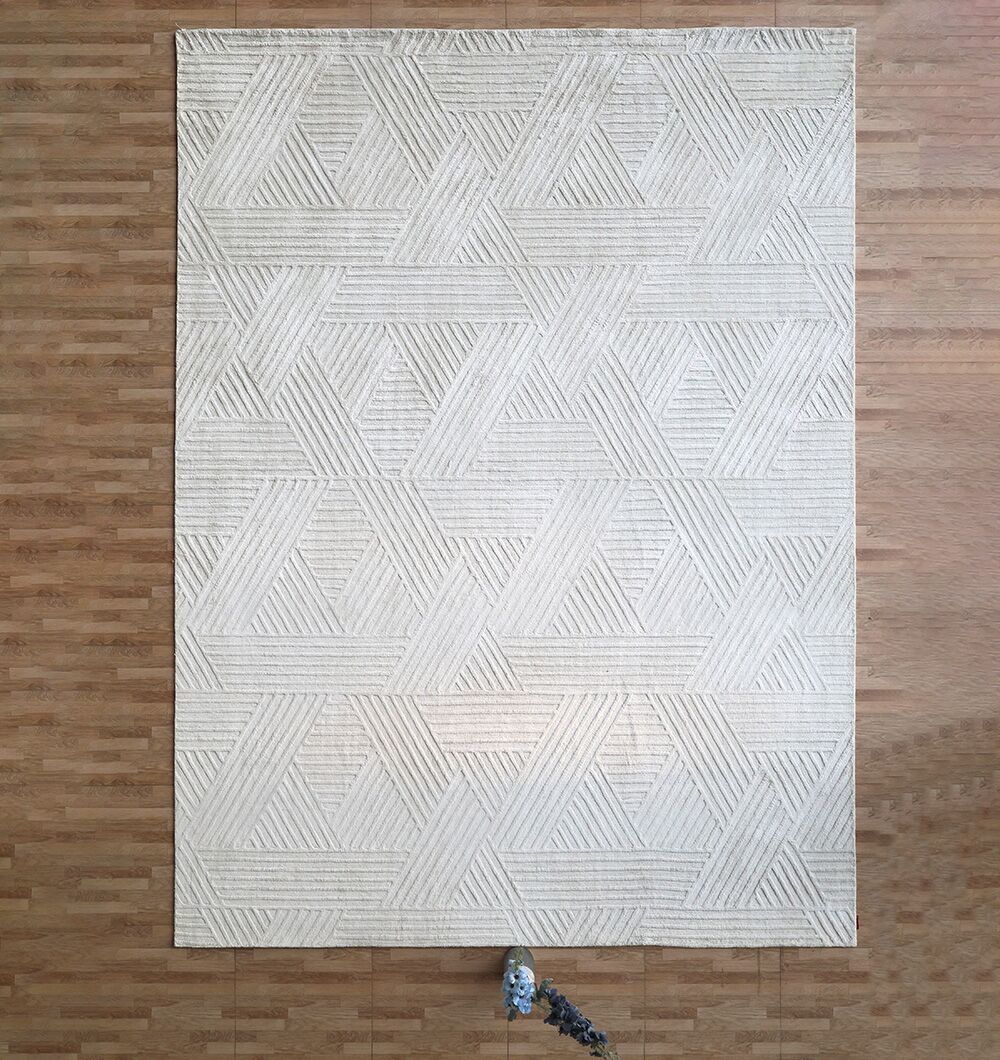 The Handmade Natural Color Area Rug - LKH 015 by RAMSHA HOME, featuring a large geometric pattern in beige with intersecting lines forming triangles, is placed on a wooden floor with a small potted plant nearby.