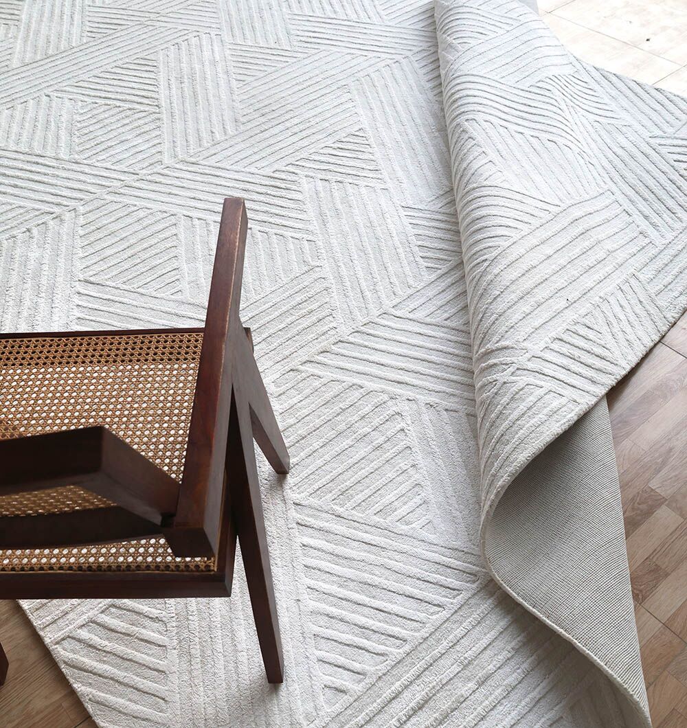 The Handmade Natural Color Area Rug - LKH 015 by RAMSHA HOME, with its textured geometric design, is partially rolled on a wooden floor. A wooden chair with a woven cane seat is placed at the rug's edge.
