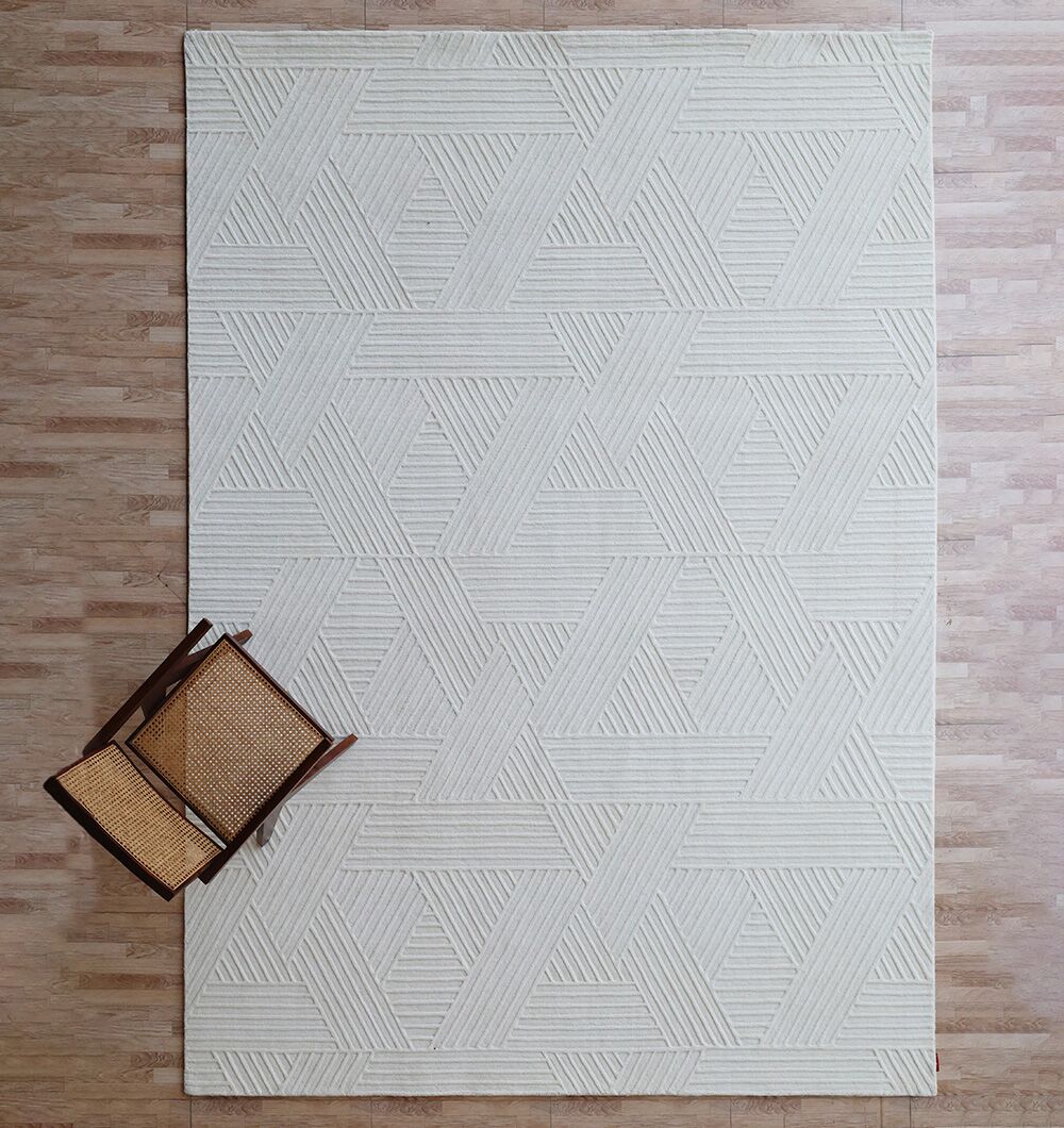 The Geometric Woven Natural Color Handmade Area Rug (LKH-016) by RAMSHA HOME lies on a light wood floor, with a wooden chair featuring a woven seat placed at the lower left corner, partially on and off the stylish ivory rug.