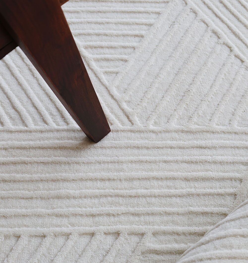 A close-up of the RAMSHA HOME Geometric Woven Natural Color Handmade Area Rug (LKH-016) shows its textured, raised stripe patterns; a dark wooden furniture leg and part of a light object are also visible.