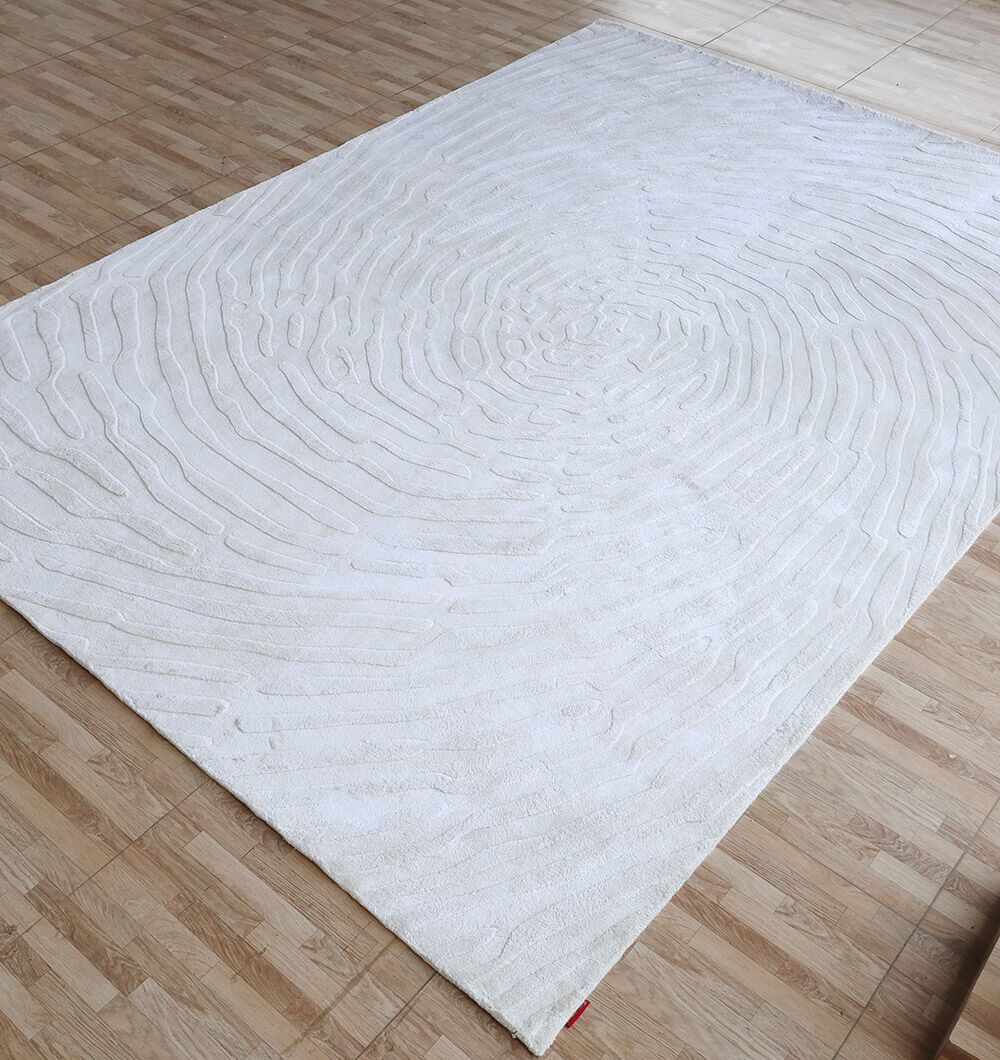 The RAMSHA HOME Decorative Handmade Area Rug - LKH-017, featuring textured concentric wave patterns in ivory, lies on a light brown wood-patterned tile floor, resembling ripples or tree rings.
