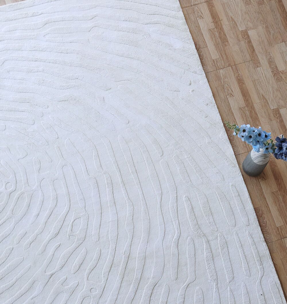 A Decorative Handmade Area Rug - LKH-017 by RAMSHA HOME with textured ivory, wavy raised patterns lies on a wooden floor, with a small vase of blue and purple flowers at its corner.