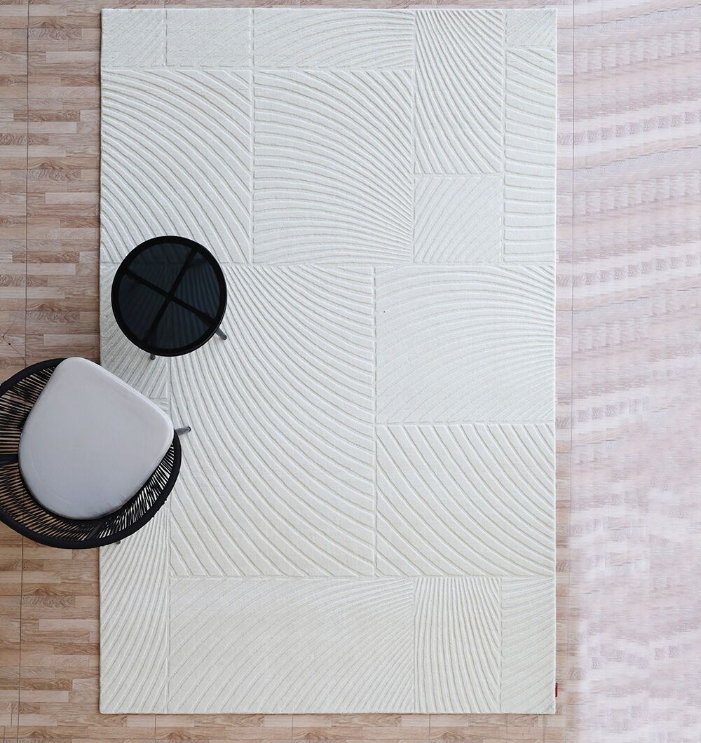 A modern living space featuring the RAMSHA HOME Natural Color Handmade Area Rug - LKH-018 (200x300 cm) in off-white with geometric and curved patterns, paired with a round black side table and a contemporary chair on a wooden floor.