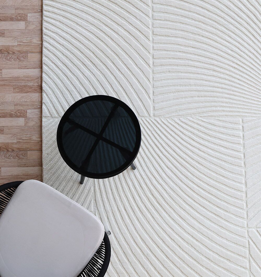 A round black side table and a modern chair sit on the RAMSHA HOME Natural Color Handmade Area Rug - LKH-018 (200x300 cm) featuring curved, geometric patterns, placed next to a light wood floor.
