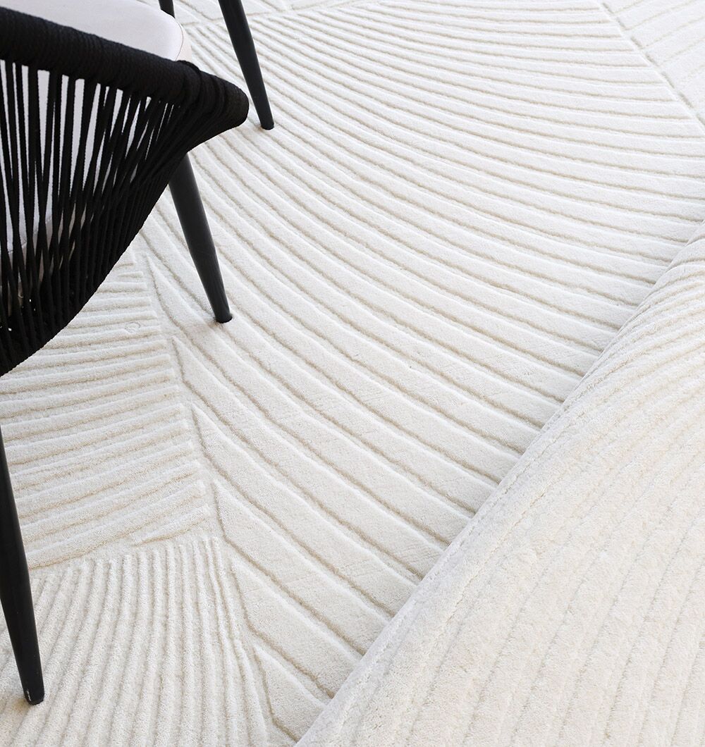 A close-up of RAMSHA HOME’s Natural Color Handmade Area Rug (LKH-018, 200x300 cm) featuring raised, wavy textures, placed beside a black chair with angled legs and a woven back.