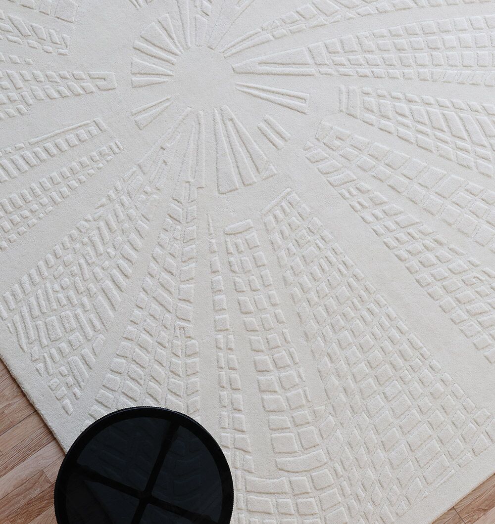 The RAMSHA HOME Handmade Decorative Area Rug - LKH-019 (200x300 cm) in ivory displays a raised sunburst pattern; a small round black table sits on one corner, revealing glimpses of the underlying wooden floor.