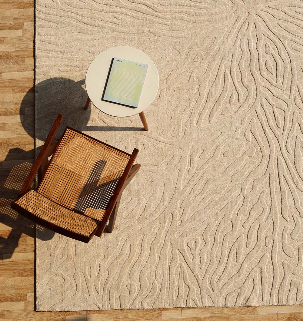 A wooden chair with a woven seat sits beside a small round table holding an open notebook, all arranged atop the RAMSHA HOME Handmade Beige Rug with Organic Tree-Inspired Pattern (LKH-025), set on a wooden floor.
