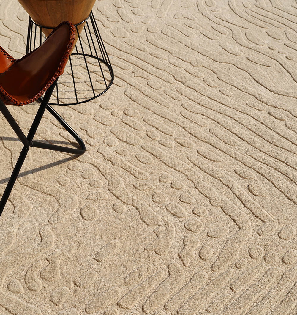The RAMSHA HOME Modern Beige Textured Area Rug – Abstract Wavy Pattern LKH-026 showcases a contemporary design with its textured beige finish and wavy, pebble-inspired patterns for a modern flair. A portion of a brown leather chair and a wooden-topped table with black metal legs can be seen in the upper left corner, complementing the inviting aesthetic of this handmade rug.