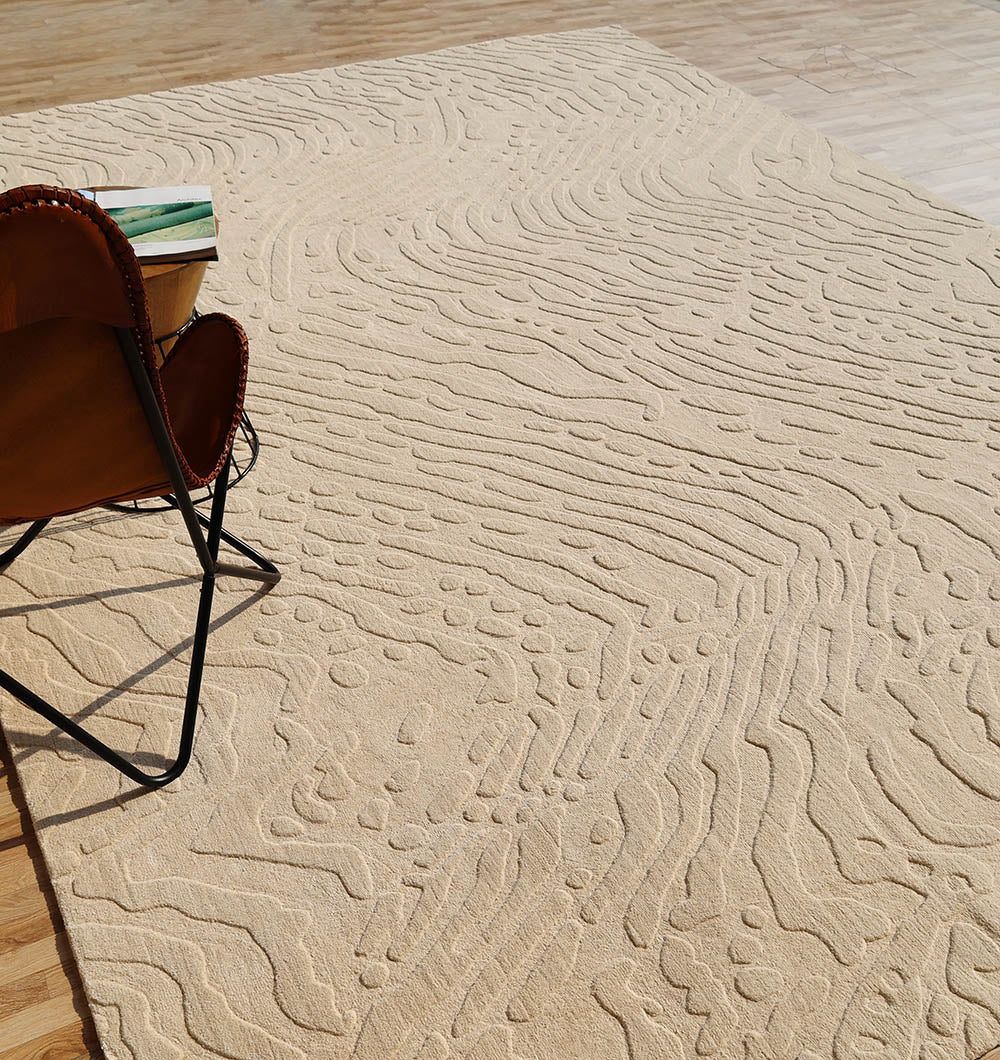A Modern Beige Textured Area Rug – Abstract Wavy Pattern LKH-026 from RAMSHA HOME, featuring raised, wavy patterns in a contemporary neutral tone, adds visual interest to the wooden floor. A brown leather chair and a book sit atop this handmade rug, creating a cozy and inviting spot for relaxation.