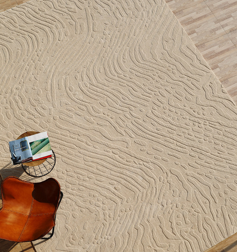 The RAMSHA HOME Modern Beige Textured Area Rug – Abstract Wavy Pattern LKH-026 lies on a wooden floor, infusing the space with a contemporary flair through its raised abstract design. In the corner, a brown leather chair and a small round table topped with a book and glasses complete the scene, as sunlight casts gentle shadows across the handmade rug and its surroundings.