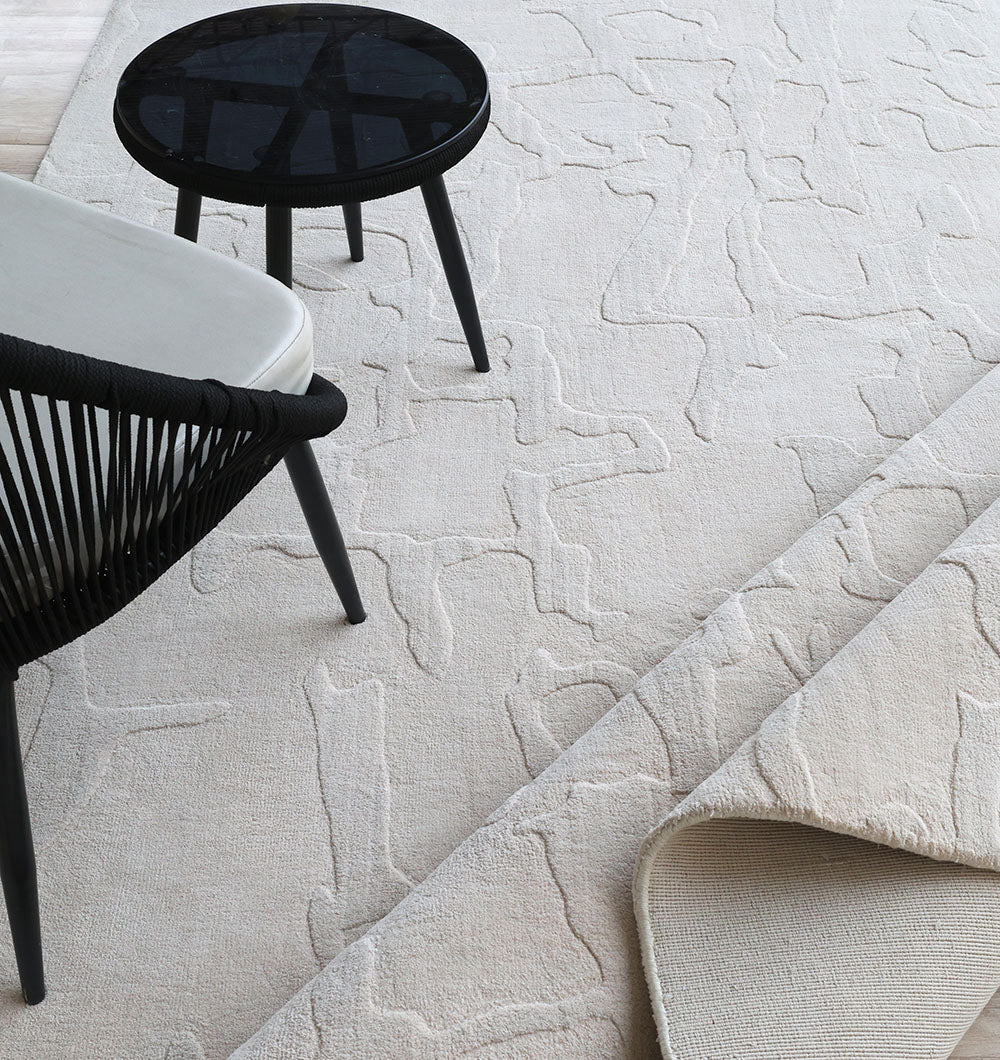 A modern black chair with woven sides and a white cushion sits next to a round black side table on the RAMSHA HOME Decorative Light Brown Color Area Rug (LKH-027, 300x400 cm), with one rug corner slightly folded.