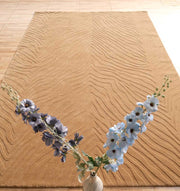 A RAMSHA HOME Decorative Handmade Beige Color Area Rug (LKH-028) lies on a tiled floor. In the foreground, a vase with light blue and purple flowers is partially visible at the image's bottom.