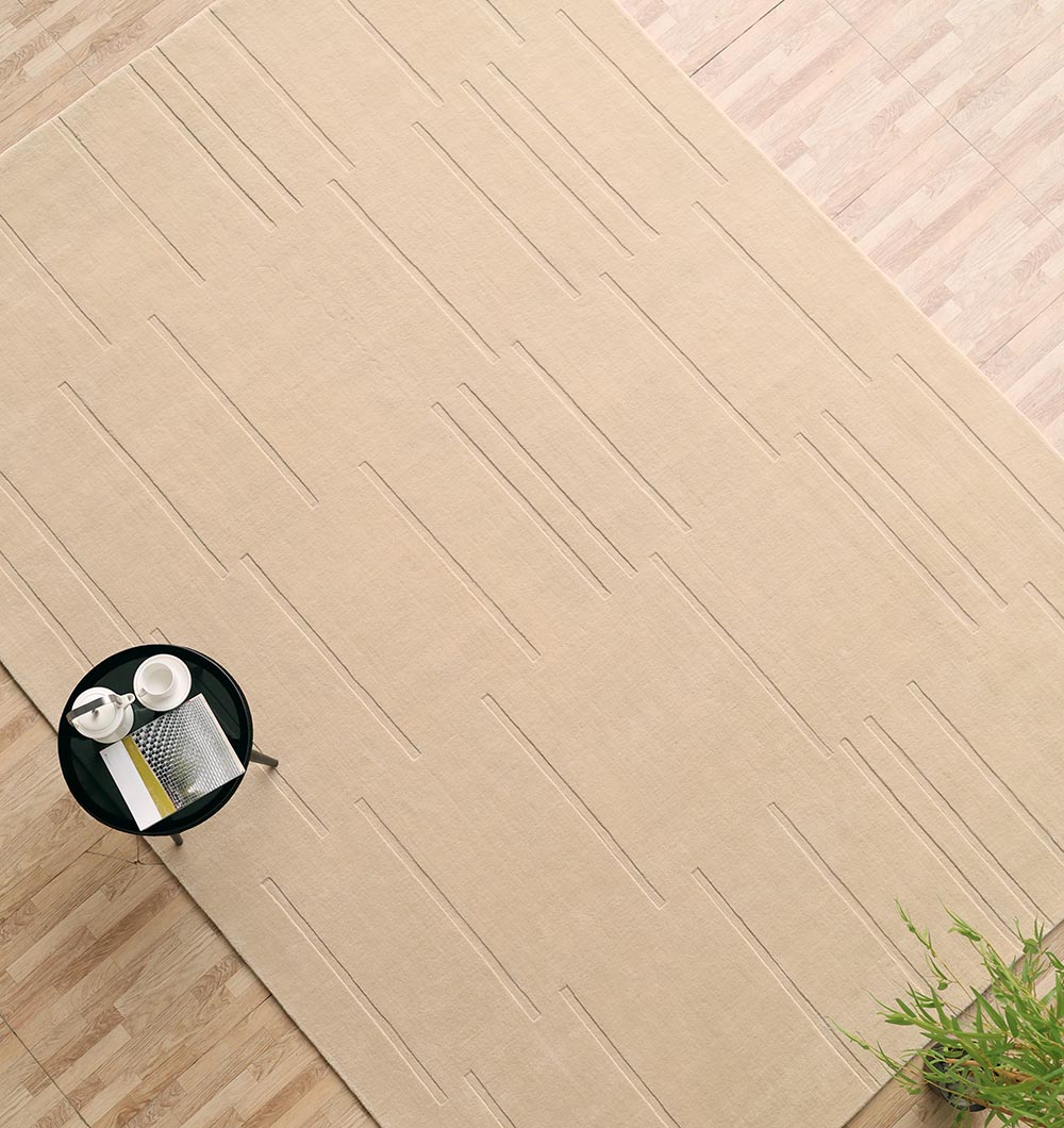 The RAMSHA HOME Rectangle Handmade Decorative Beige Area Rug (250x350 cm, LKH-031) lies on a light wooden floor, topped with a small round black table holding a book, cup, and teapot; a green plant adds a fresh touch to the corner.