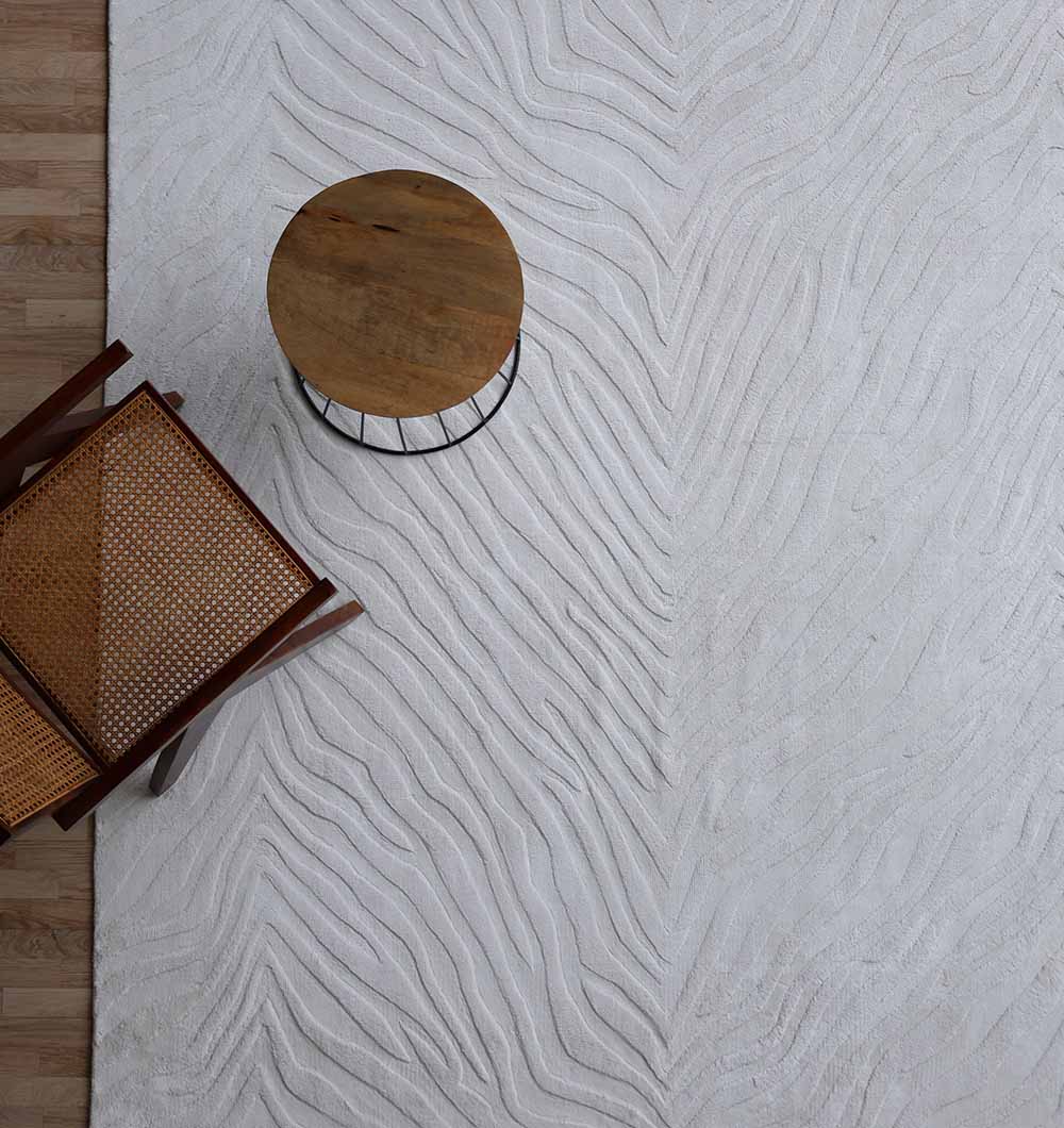 A woven chair and round wooden side table rest on the Rectangle Handmade Natural Color 200x300 cm area rug by RAMSHA HOME, featuring wavy, leaf-like patterns—an ideal modern accent atop a light wood floor.