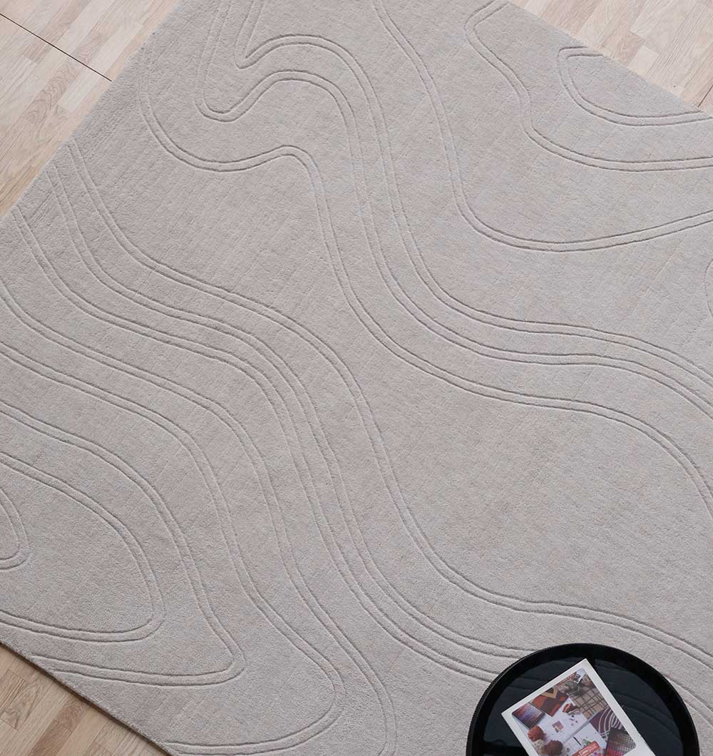 The RAMSHA HOME Rectangle Handmade Lt.Grey Color 200x300 cm Area Rug (LKH-036) with raised, wavy patterns lies on a light wooden floor, while a round black table in the corner holds a magazine and a glass.