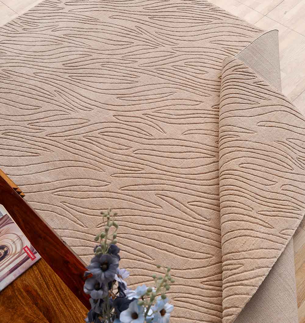 The RAMSHA HOME Rectangle Handmade Beige 200x300 cm Area Rug (LKH-042) is shown partially folded on a light wood floor, beside a wooden coffee table, magazines, and a vase with purple flowers for a cozy modern look.