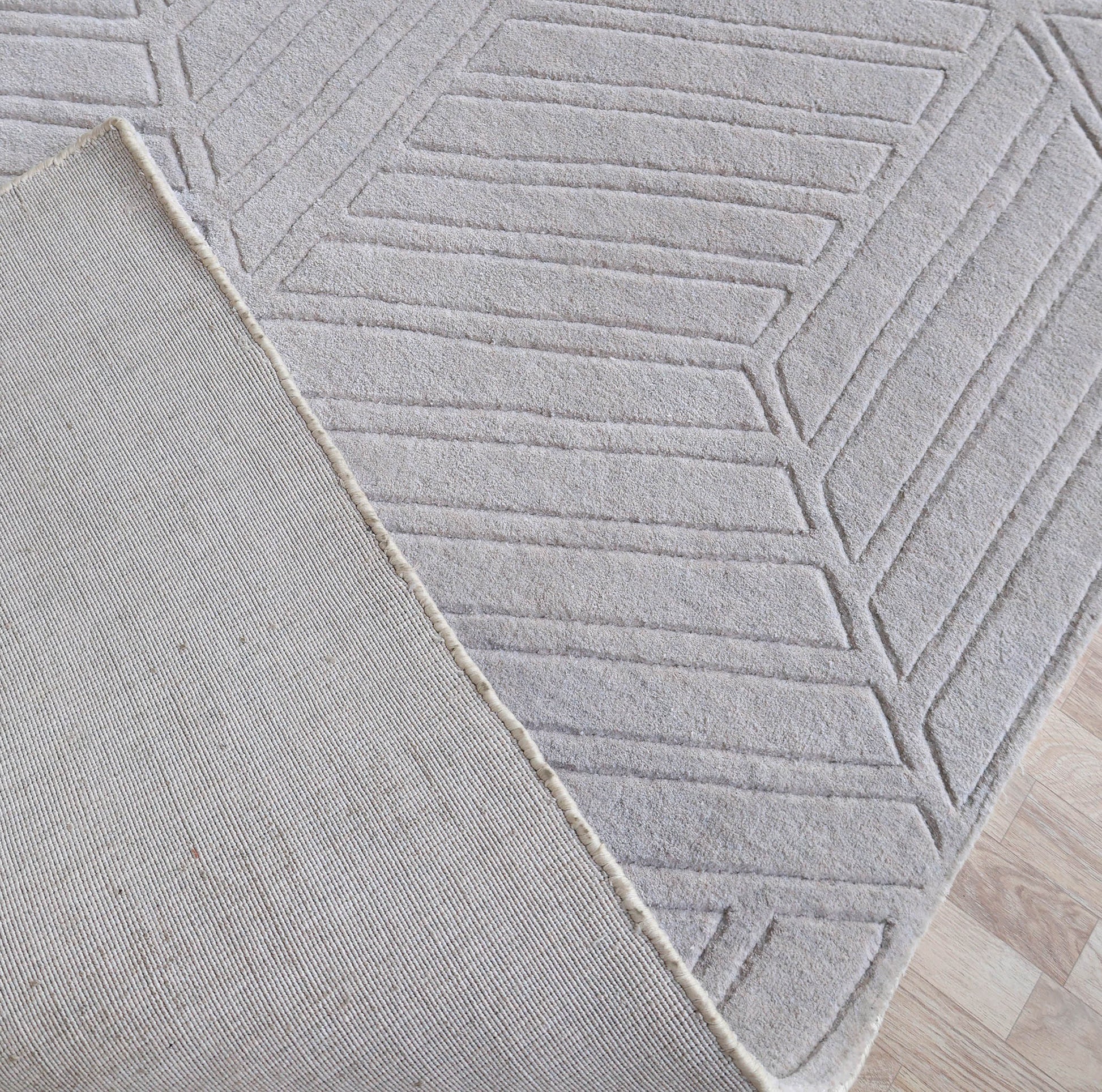 A close-up of the RAMSHA HOME Home Decor Light Grey Handmade Area Rug (300x400 cm, LKH-044) shows a modern geometric pattern; one corner is flipped to reveal the plain underside, and the rug rests on a light wood floor.