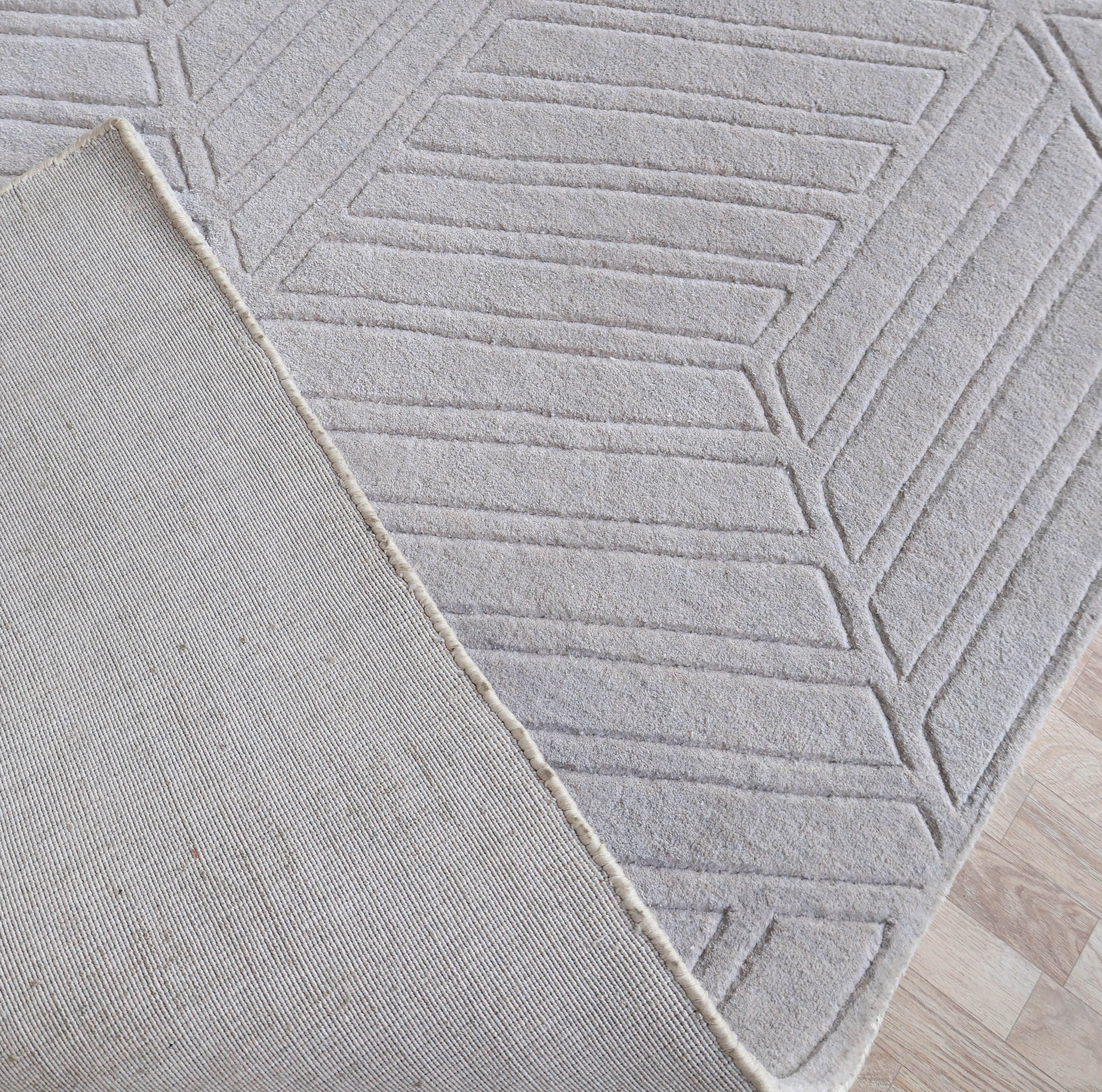A close-up of the RAMSHA HOME Home Decor Light Grey Handmade Area Rug (300x400 cm, LKH-044) shows a modern geometric pattern; one corner is flipped to reveal the plain underside, and the rug rests on a light wood floor.
