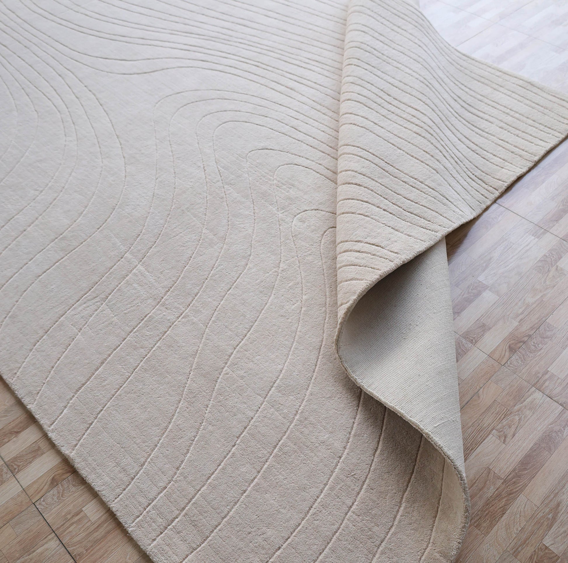 The RAMSHA HOME Modern Handmade Area Cream Color Rug (300x400 cm - LKH-046) features textured, wavy raised lines and soft, woven material, shown partially folded to reveal its quality craftsmanship and cream hue on a light wood floor.