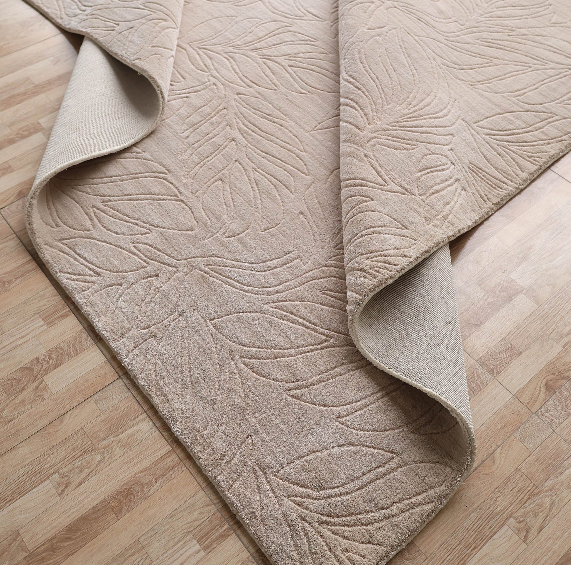 The RAMSHA HOME Nature Leaf Handmade Contemporary Rug (250x350 cm, LKH-048) features a neutral ivory tone with a subtle leaf pattern, shown partially folded on a light wood floor to display its textured top and plain underside.