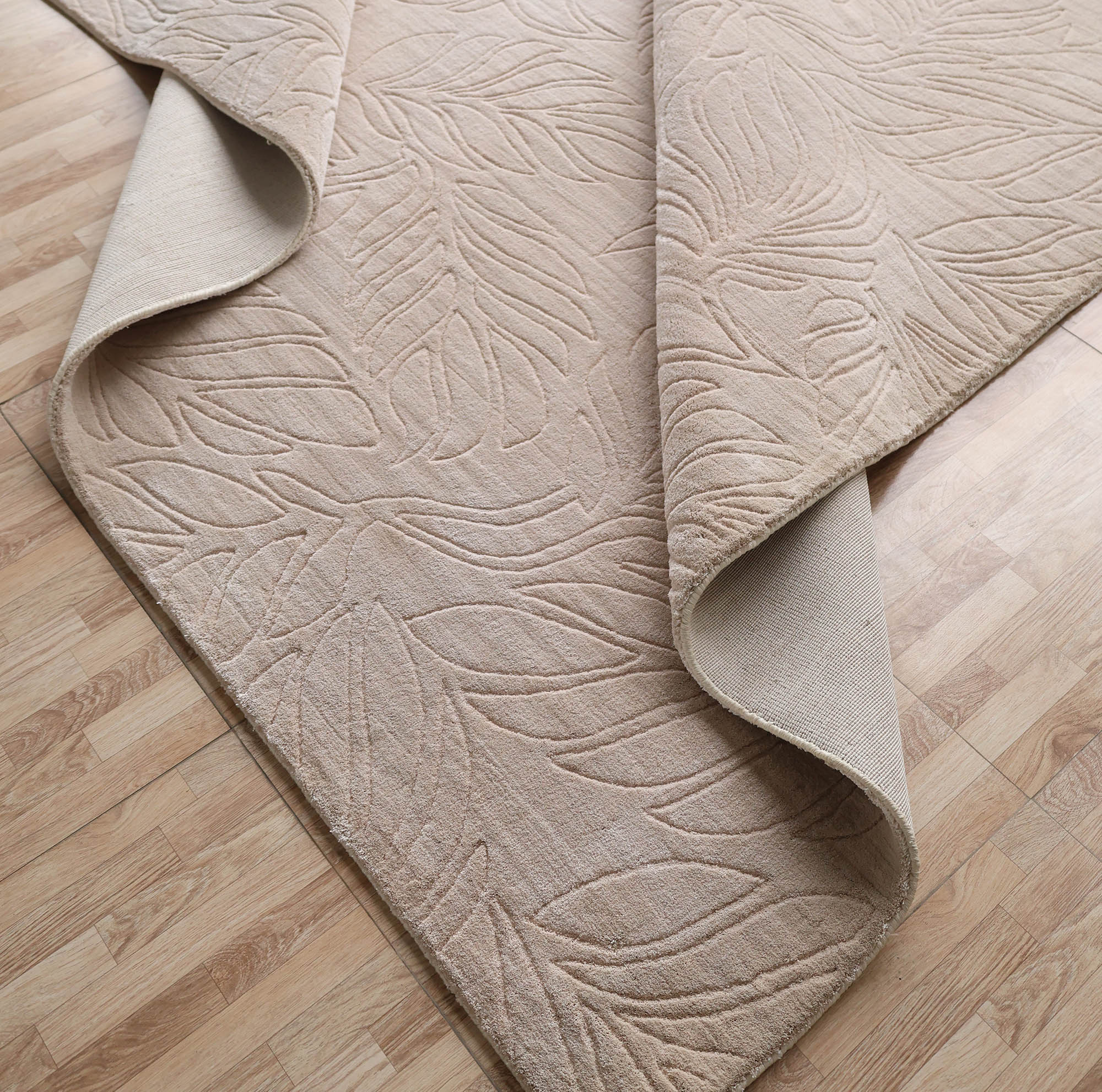 The RAMSHA HOME Nature Leaf Handmade Contemporary Rug (250x350 cm, LKH-048) features a neutral ivory tone with a subtle leaf pattern, shown partially folded on a light wood floor to display its textured top and plain underside.