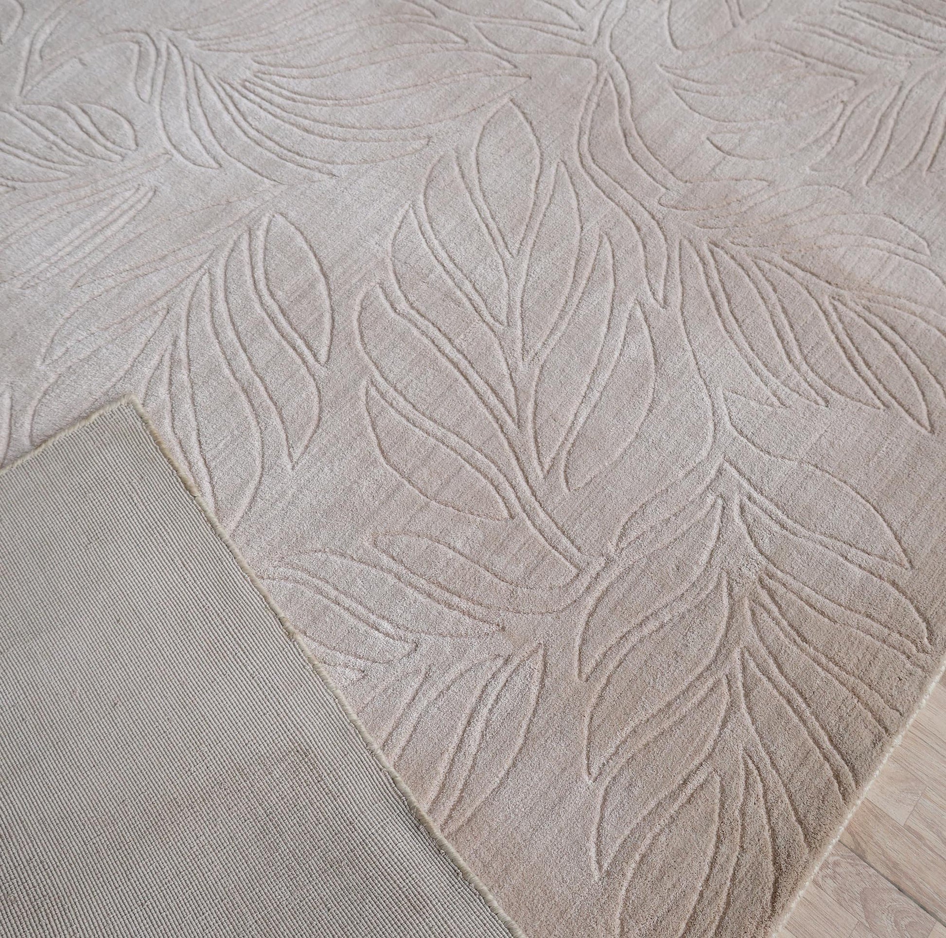 A close-up of the RAMSHA HOME Nature Leaf Handmade Contemporary Rug 250x350 cm (LKH-048) in neutral ivory, featuring a raised botanical leaf pattern, partially flipped to reveal its plain underside and textured design on a light wooden floor.