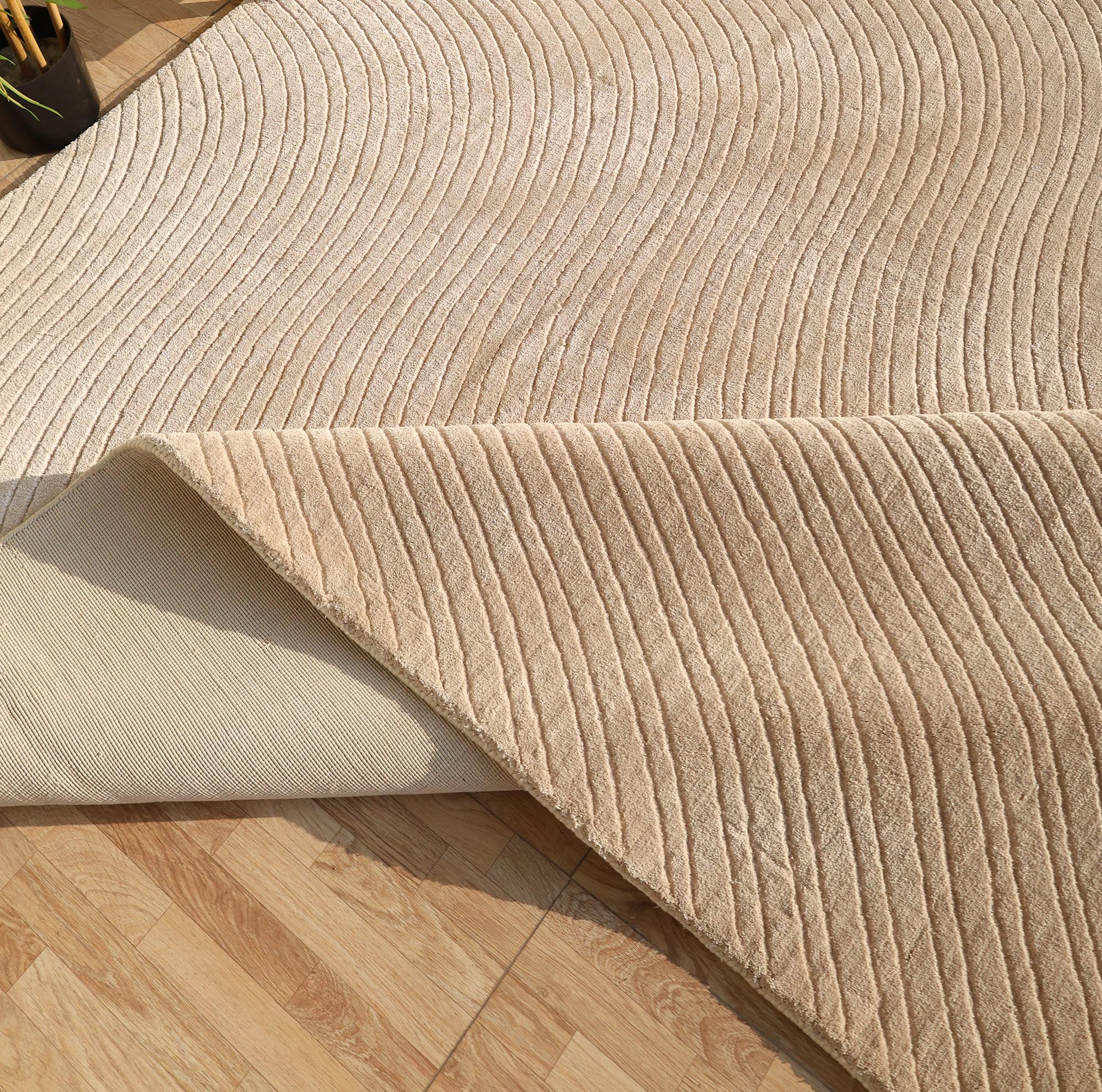 The RAMSHA HOME Handmade beige Wave Modern Abstract High-Low Area Rug (250x350 cm, LKH-049) is shown partially folded to reveal its underside on a light wood floor, with sunlight and a plant in the upper left corner.