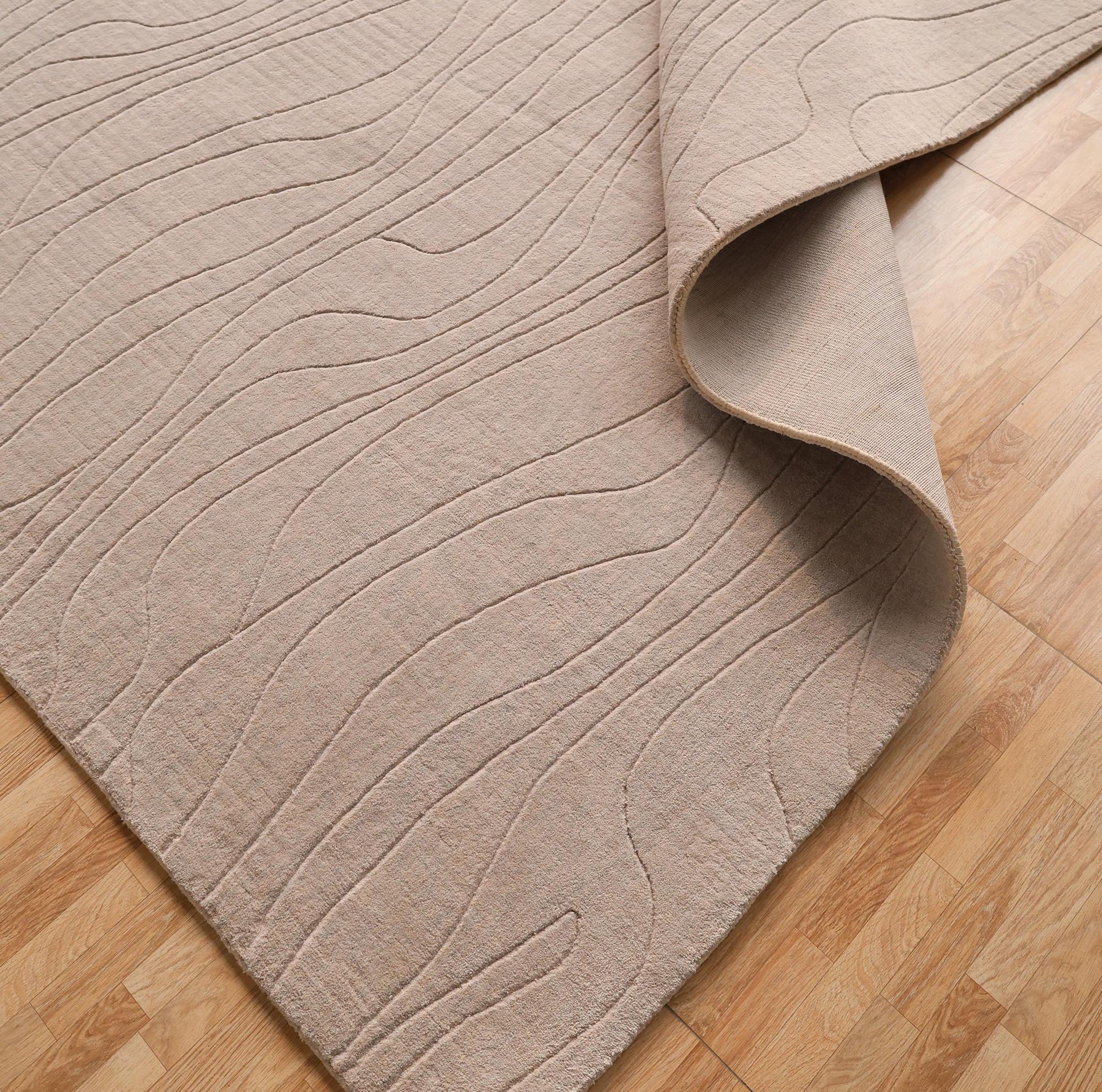 The RAMSHA HOME Light Grey Color Handmade 250x350 cm Area Rug (LKH-043) features subtle wavy embroidery and a textured, minimalist look, shown partially folded on a light wood floor.