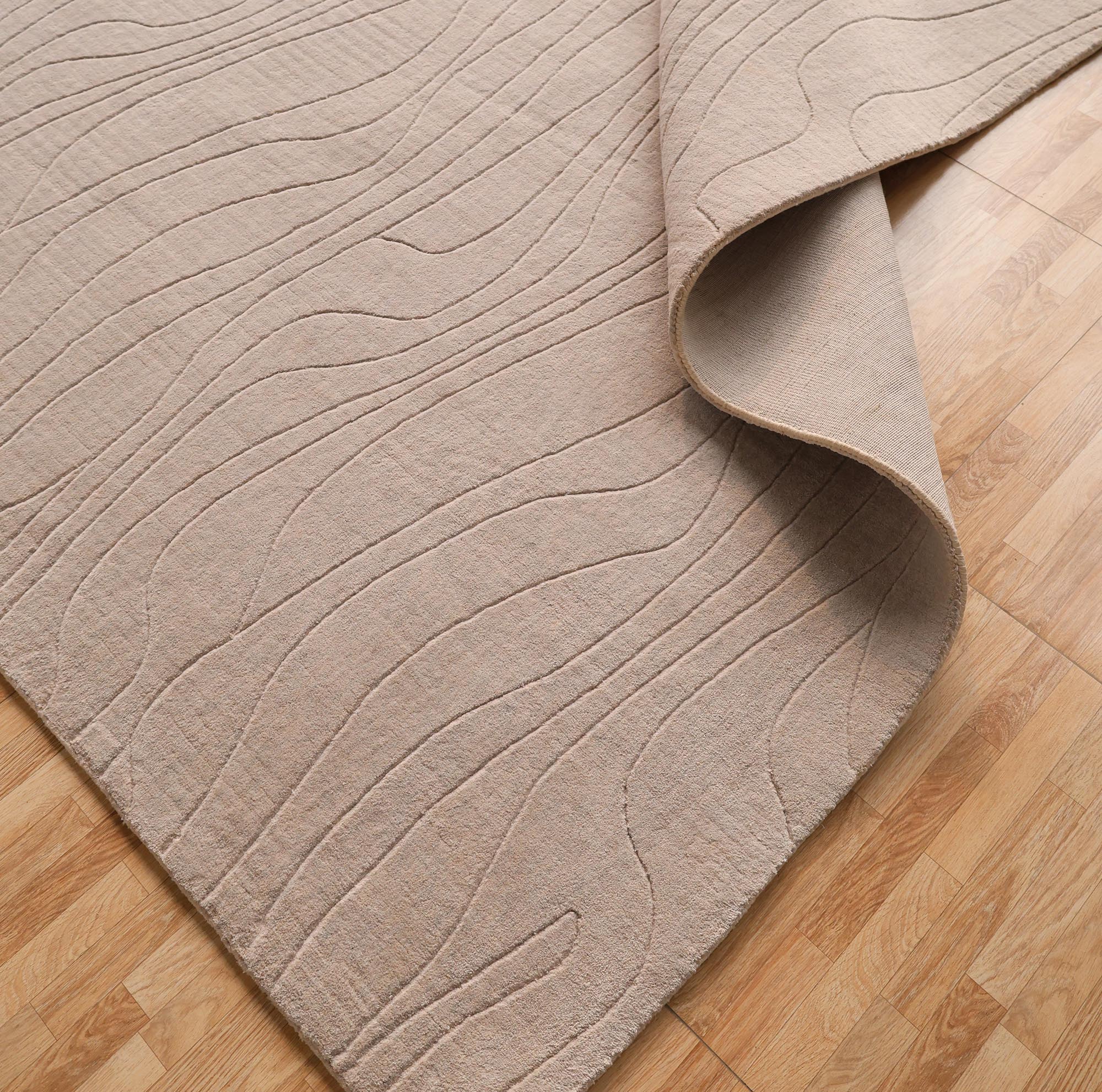 The RAMSHA HOME Light Grey Color Handmade 250x350 cm Area Rug (LKH-043) features subtle wavy embroidery and a textured, minimalist look, shown partially folded on a light wood floor.