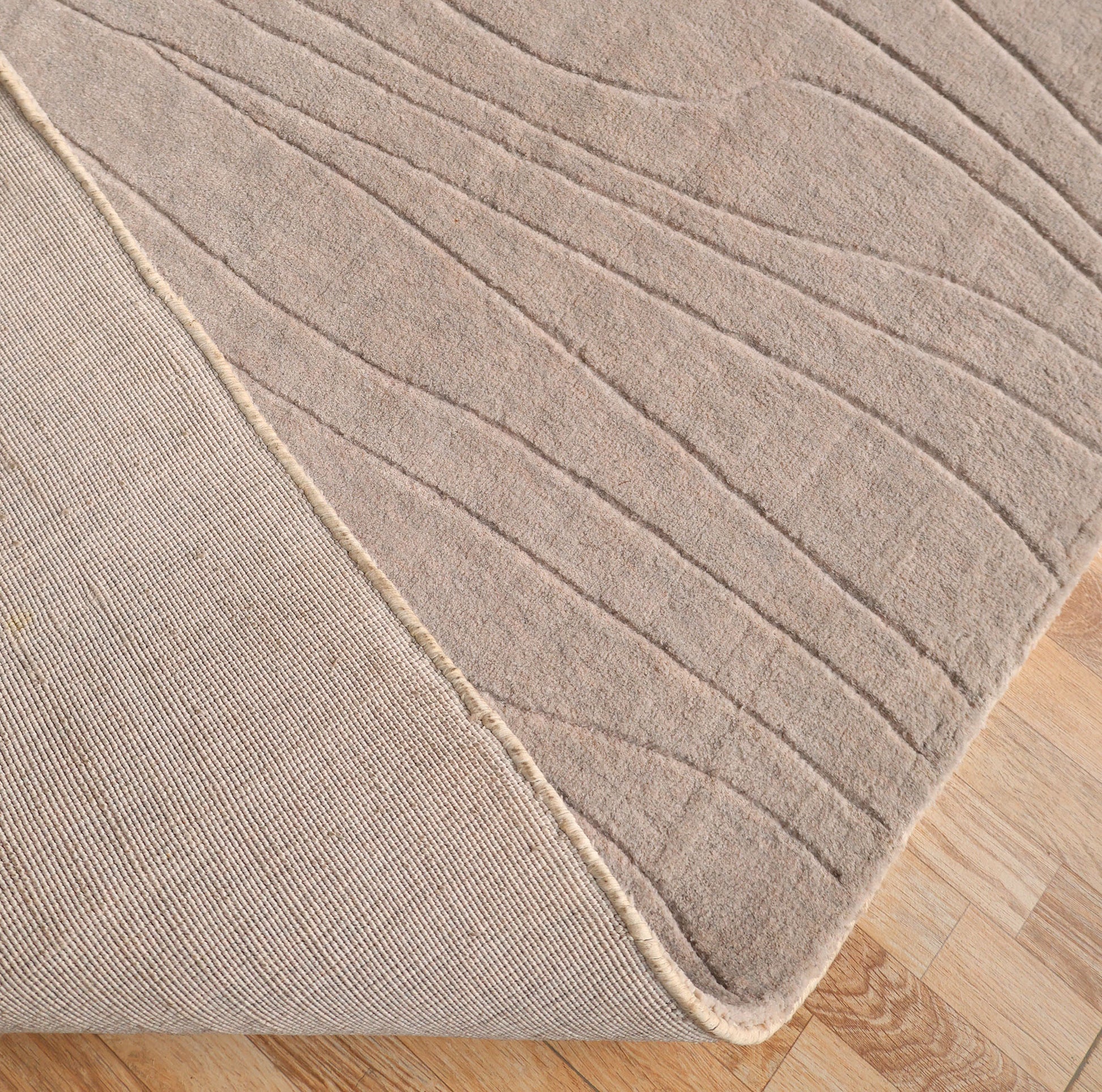 A close-up of the RAMSHA HOME Light Grey Color Handmade 250x350 cm Area Rug (LKH-043) on a wooden floor, with its corner folded to display the textured underside and subtle wavy line patterns.