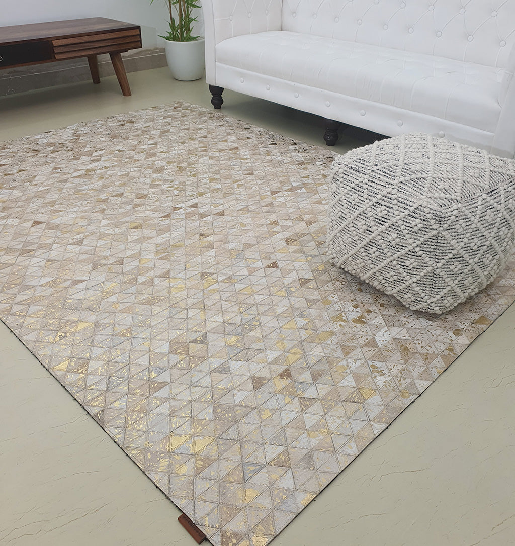 A modern living room with a white tufted sofa, the Amazing Grey & Tan Color Playroom Leather Rug - LR 006 featuring gold accents, a textured round pouf, a wooden coffee table, and a potted plant in the corner.