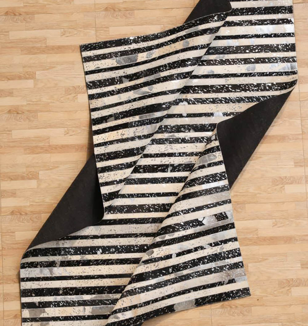 The RAMSHA HOME Striped Abstract Black & Ivory Leather Area Rug LR-009, featuring a distressed splatter design, is partially folded on a light wood floor for a modern touch.