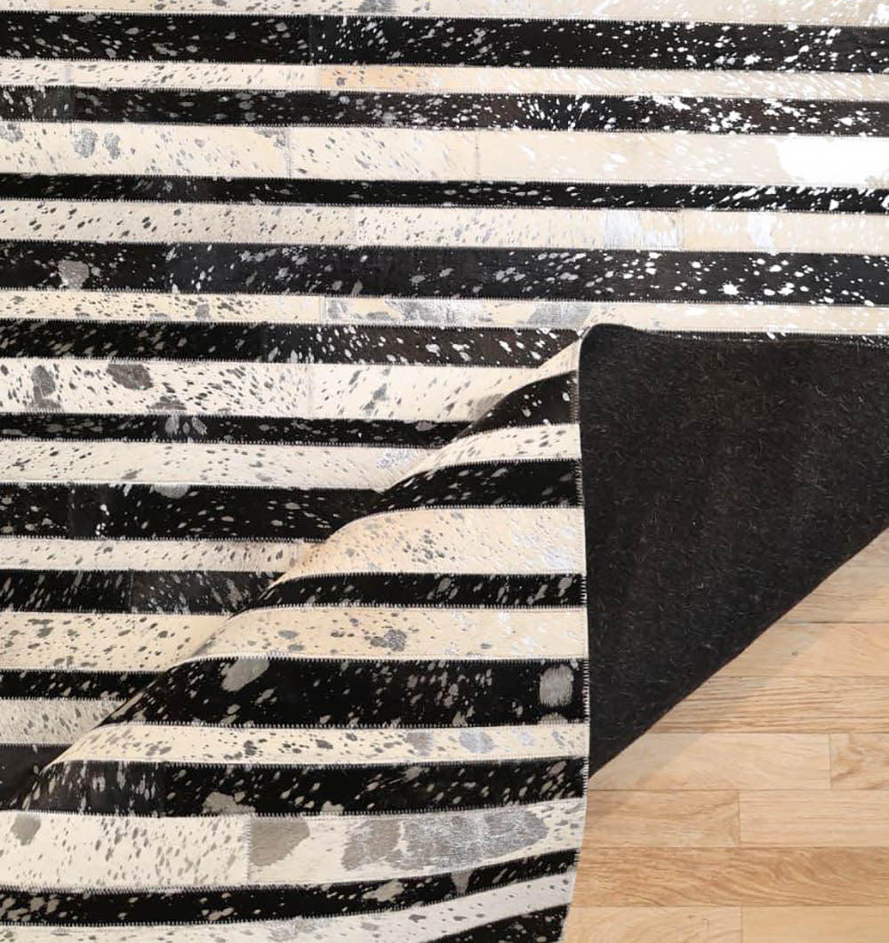The RAMSHA HOME Striped Abstract Black & Ivory Leather Area Rug LR-009, featuring irregular speckled patterns, is shown partially folded at one corner on a light wooden floor.