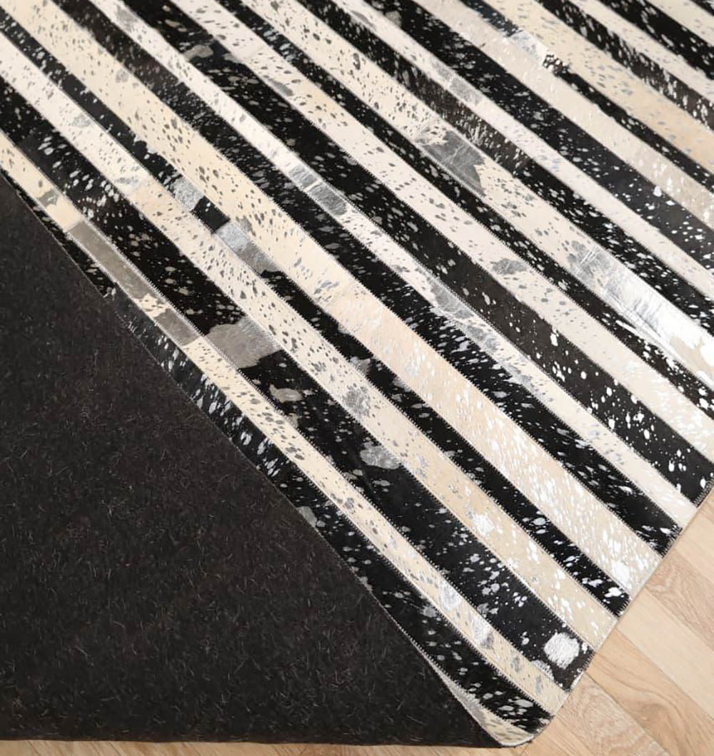 A close-up of the RAMSHA HOME Striped Abstract Black & Ivory Leather Area Rug LR-009 shows black, white, and silver diagonal stripes, with a corner folded to reveal its solid dark underside and wooden floor below.