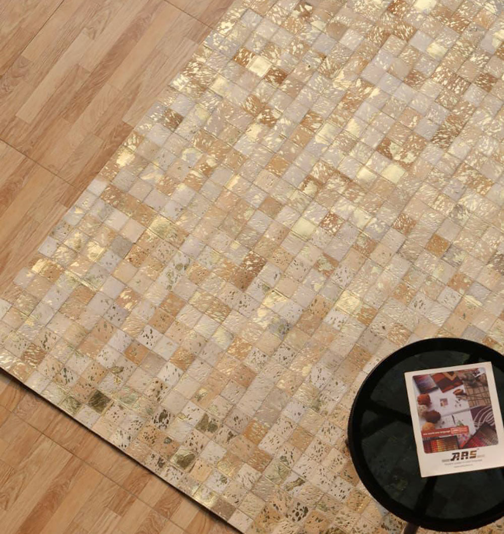 A close-up of the Handmade Mosaic Patchwork Beige & Tan Leather Rug LR-015 by RAMSHA HOME, featuring a square tile pattern on a wooden floor. A round black table with a magazine is partly visible in the lower right corner.