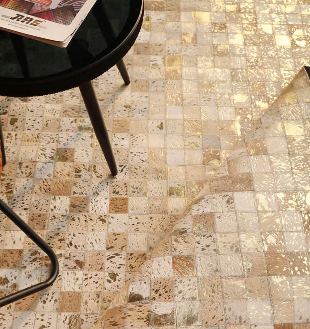 A close-up of a gold and silver mosaic-tiled floor with a protective sheet partially peeled back. A round black table with slender legs and a magazine sits in the corner beside the RAMSHA HOME Handmade Mosaic Patchwork Beige & Tan Leather Rug LR-015.