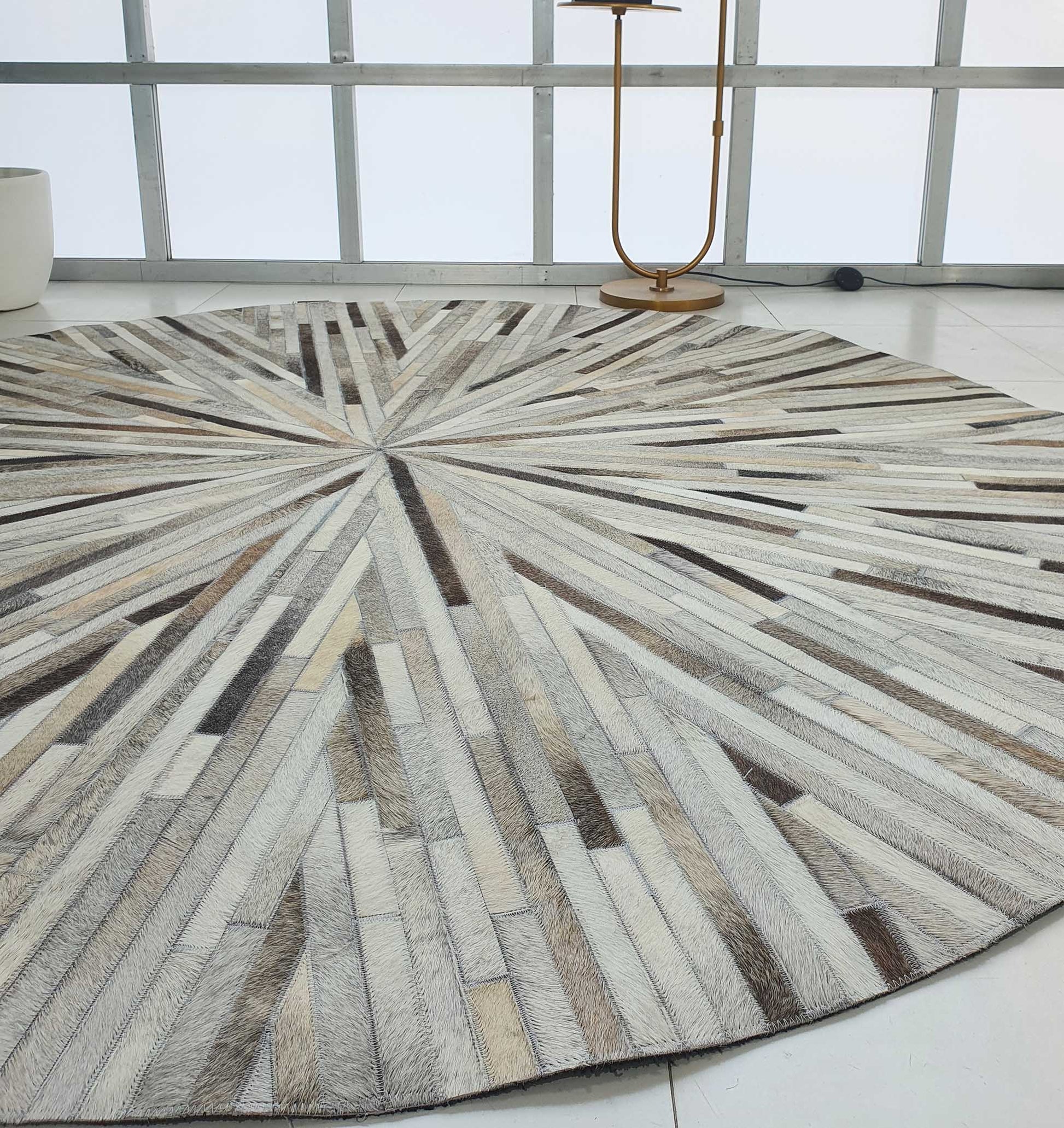 The RAMSHA HOME Home Decorative Leather Rug - LR 018 features a round starburst pattern in beige, brown, and gray segments, shown on a white tiled floor in a modern room with large windows and a gold floor lamp.